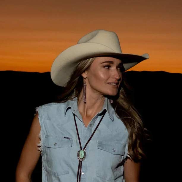 Bri Bagwell wearing a cowboy hat and denim vest against an orange sunset sky.
