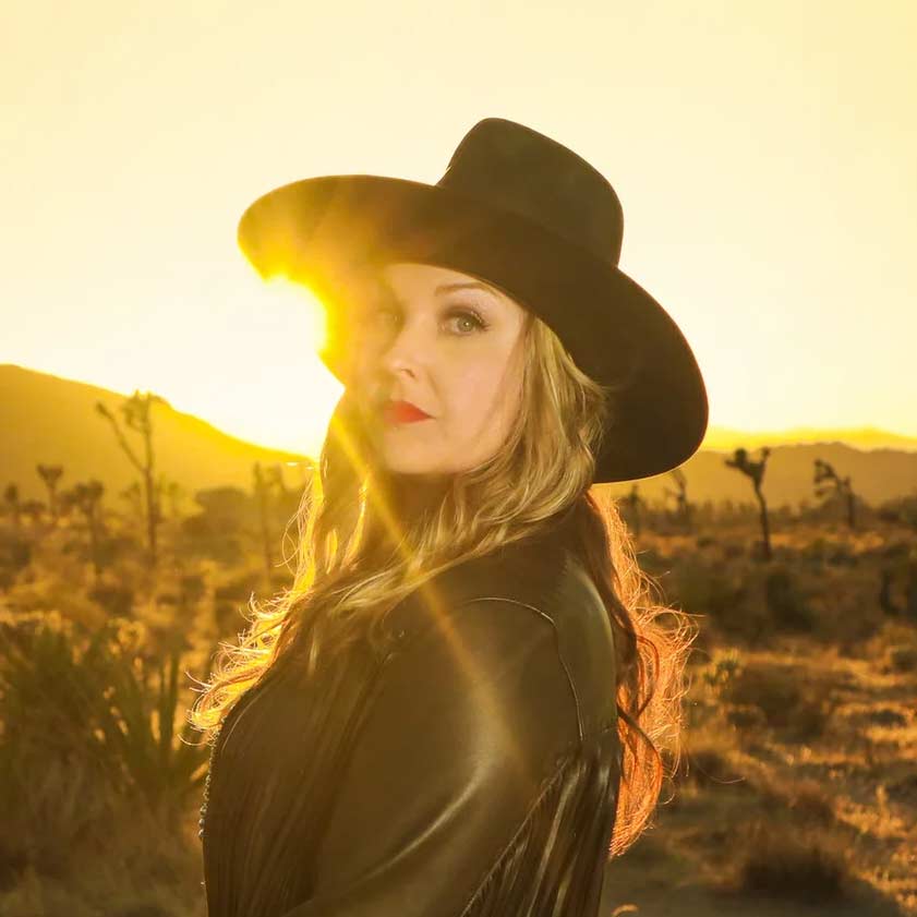 Sunny Sweeney wearing a black cowboy hat in a desert landscape with a sunset.